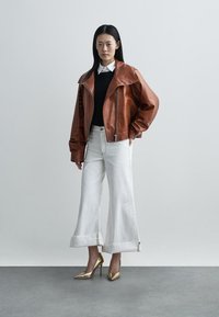 Brown leather jacket with wide lapels, black sweater, and white wide-leg jeans. Gold high heels complete the outfit. Simple, minimalist design.