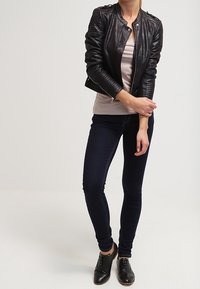 Kobieta w czarnej skórzanej kurtce, beżowej bluzce, ciemnych obcisłych dżinsach i czarnych sznurowanych butach, stoi z założonymi rękami na biodrach.