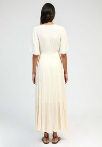 Robe maxi en tissu crème clair, avec des manches courtes et flottantes, une taille ajustée et une jupe à volants. Texture lisse avec un subtil éclat.