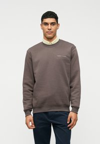 Uomo che indossa un maglione a girocollo marrone sopra una camicia beige con bottoni e pantaloni scuri, in piedi contro uno sfondo bianco semplice.