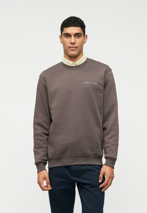 Bărbat îmbrăcat cu un pulover cu guler rotund maro peste o cămașă bej cu nasturi și pantaloni negri, stând în fața unui fundal alb simplu.