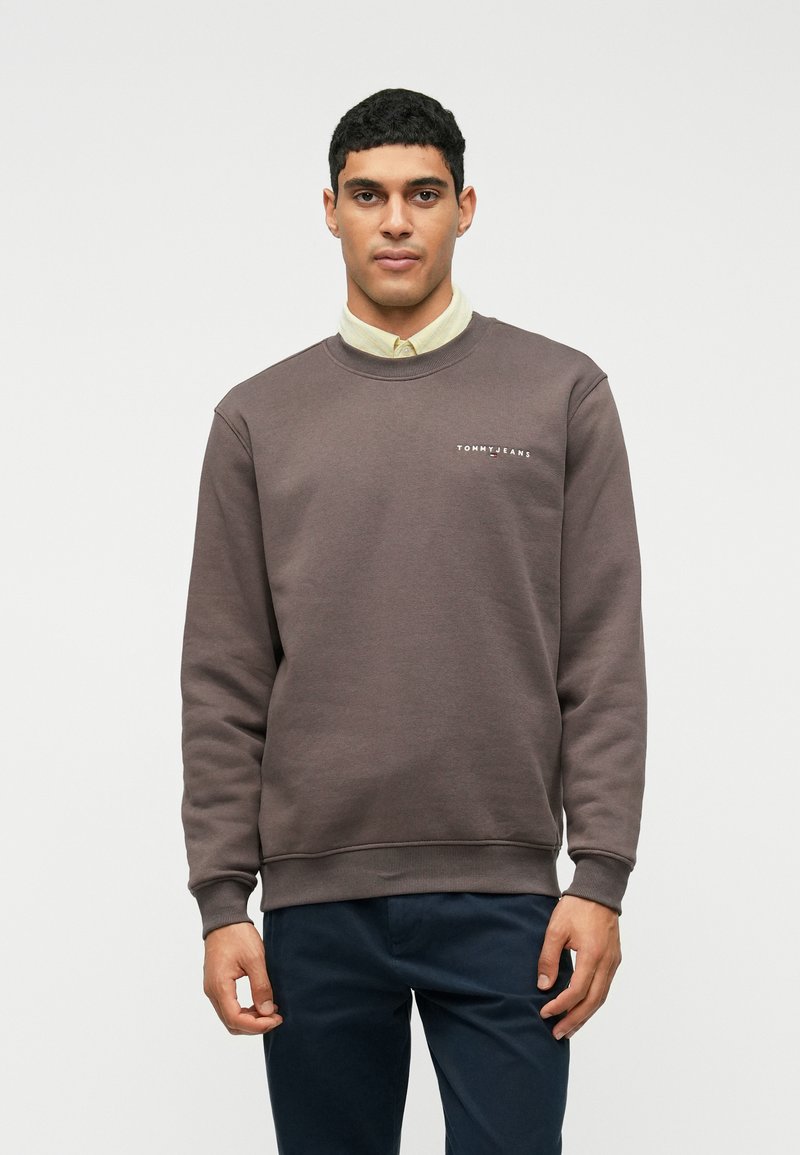 Uomo che indossa un maglione a girocollo marrone sopra una camicia beige con bottoni e pantaloni scuri, in piedi contro uno sfondo bianco semplice.