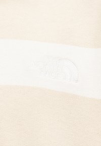 Κοντινή λήψη μπεζ υφάσματος με λευκή οριζόντια ρίγα και διακριτικό λευκό κεντημένο λογότυπο «The North Face».