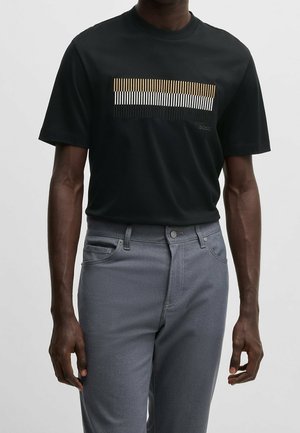 Man draagt een zwart T-shirt met korte mouwen met horizontale gouden, witte en zwarte strepen en een grijze broek tegen een effen achtergrond.