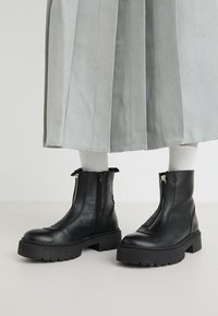 Botas tobilleras de cuero negro con suela gruesa, con una cremallera frontal y detalles de costura. Acompañadas de calcetines blancos acanalados y una falda clara.