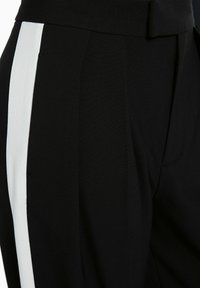 Pantalon noir cintré avec une taille haute et une bande blanche verticale le long de la couture latérale.