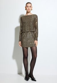 Robe mini dorée pailletée avec manches longues et coupe ample, dotée d'un col rond et d'une taille cintrée. Collants noirs et chaussures à bout pointu.