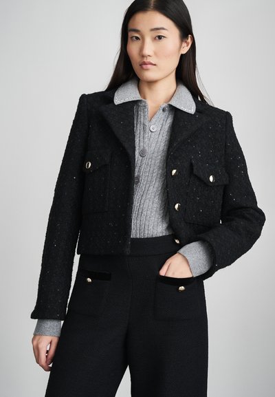 Veste noire cropped en tissu texturé, avec de grandes poches avant et des boutons dorés. Portée sur un pull gris à côtes et un pantalon noir ajusté.