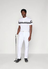 Vitt pikétröja med svarta horisontella ränder, kombinerad med vita byxor och svarta skor; håller i en golfklubba. Slät textur, atletisk passform.