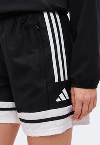 Μαύρο και άσπρο σορτς Adidas με τρεις λευκές ρίγες στο πλάι και το λογότυπο Adidas κοντά στο τελείωμα, φορεμένο με μαύρο μπουφάν.