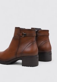 Botas de tobillo de cuero marrón con punta redondeada, tacón bajo cuadrado, cremallera lateral y una correa ajustable con un detalle de hebilla de metal.