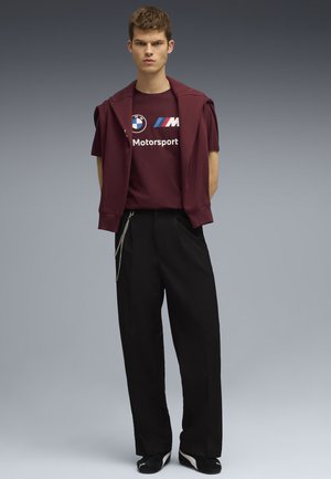 Bordo majica kratkih rukava s BMW Motorsport logotipom, složena ispod bordo prsluka bez rukava, u kombinaciji s crnim širokim hlačama i crnim cipelama.