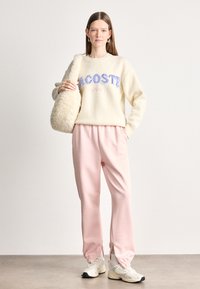 Κρέμα πουλόβερ με υφή και μπλε και ροζ γράμματα, συνδυασμένο με απαλό ροζ παντελόνι που διαθέτει ελαστικά και φερμουάρ. Το μοντέλο κρατά μια αφράτη τσάντα.