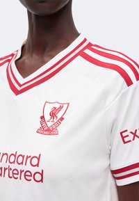 Λευκή φανέλα της Liverpool Football Club με κόκκινες ρίγες στους ώμους και το γιακά, με το έμβλημα του συλλόγου και τα λογότυπα των χορηγών.