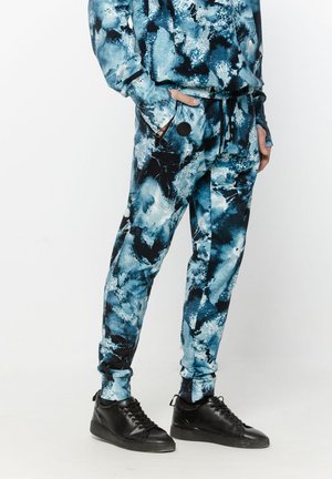 Osoba w joggerach i dopasowanym topie w niebiesko-czarny abstrakcyjny wzór, w czarnych trampkach, z rękami w kieszeniach, stojąca na białym tle.