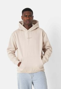 Felpa beige con tasca a marsupio e cappuccio con cordino regolabile. Presenta un piccolo logo sul petto e polsini a coste. Abbinata a jeans azzurri chiari.