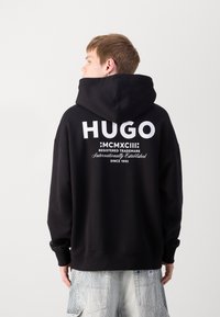 Schwarzer Kapuzenpullover mit großem weißen "HUGO"-Logo auf dem Rücken, ausgestattet mit einer Kordelzugkapuze, gerippten Bündchen und einem lässigen Schnitt.