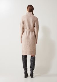 Beige pletené šaty s vysokým golierom, zúženým pásom a dlhými rukávmi. Obsahujú rebrovaný lem a opasok. Nosené s čiernymi čižmami po kolená.