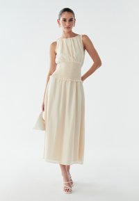 Robe midi couleur crème avec une taille smockée et un tissu froncé. Matière texturée et légère avec une jupe fluide, associée à des sandales à brides.
