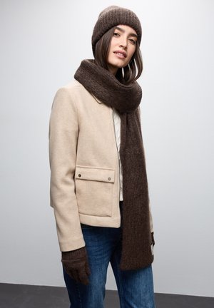 Cappotto di lana beige con bottoni argento, berretto di maglia marrone, sciarpa abbinata e guanti scuri, indossato con jeans blu su uno sfondo neutro.