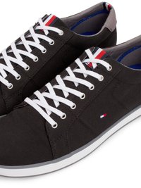 Sapatilhas de lona pretas com atacadores brancos, apresentando uma sola de borracha e um detalhe em vermelho, branco e azul no calcanhar. Logótipo discreto na lateral.