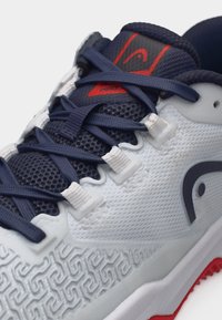 Weißer und marineblauer Sportschuh mit gemustertem Seitenteil, marineblauen Schnürsenkeln, die durch weiße Ösen gefädelt sind, und roten Details an Zunge und Sohle.