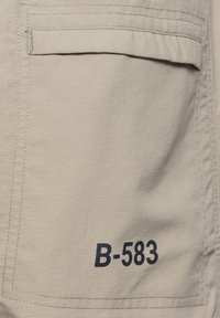 Close-up van een beige stoffen zak met klep en zwarte bedrukte tekst "B-583" in de rechteronderhoek.