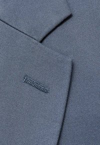 Dunkelblauer Stoff mit einer strukturierten Webart, der überlappende Bereiche und ein kleines gesticktes Detail auf der rechten Seite aufweist.