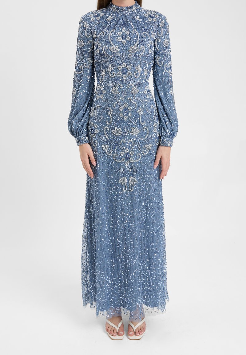 Robe bleue longue à manches longues avec une broderie complexe de perles et de sequins argentés, portée avec des sandales blanches à brides sur un fond uni.