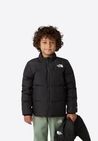 Černá péřová bunda s vysokým límcem a zapínáním na zip, s logem na hrudi, kterou drží dítě oblečené v zelených kalhotách.