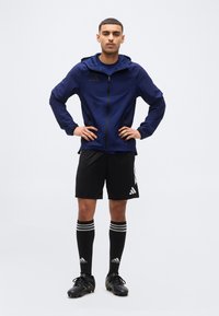 Νέος άνδρας με ναυτικό μπλε μπουφάν με φερμουάρ, μαύρα αθλητικά σορτς, μαύρες κάλτσες Adidas μέχρι τα γόνατα και μαύρα ποδοσφαιρικά παπούτσια, στέκεται με τα χέρια στη μέση.