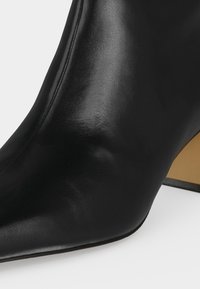 Bottine en cuir noir lisse à bout pointu avec talon bloc incliné couleur beige sur fond blanc.