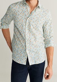 Mann in einem hellblauen, floral gemusterten Langarmhemd mit hochgekrempelten Ärmeln und dunkelblauen Jeans, der mit einer Hand hinter dem Rücken steht.