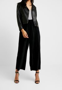 Giacca in pelle nera, top grigio, pantaloni neri a vita alta plissettati e sandali neri con cinturini alla caviglia. Tessuti lisci e design aderente.