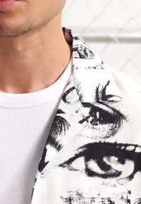 Weißes Hemd mit gerippter Textur, kombiniert mit einer gemusterten Jacke, die schwarz-weiße Augendarstellungen auf glattem Stoff zeigt.