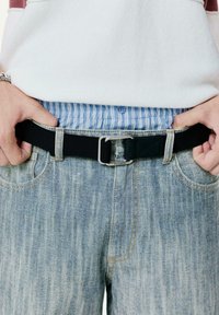 Curea neagră cu o cataramă metalică argintie, purtată peste blugi decolorati. O cămașă cu dungi albastre, deschise, este vizibilă sub talia.