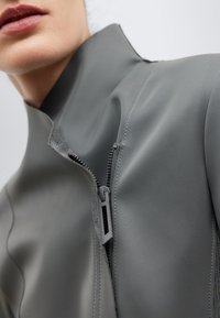 Giacca grigia con colletto alto, dotata di chiusura con zip. Il materiale ha una texture sintetica liscia e presenta cuciture visibili.