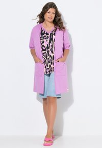 Lichtpaarse cardigan met korte mouwen en twee voorzakken, over een zwart-roze luipaardprint top, gecombineerd met denim shorts en roze sandalen.