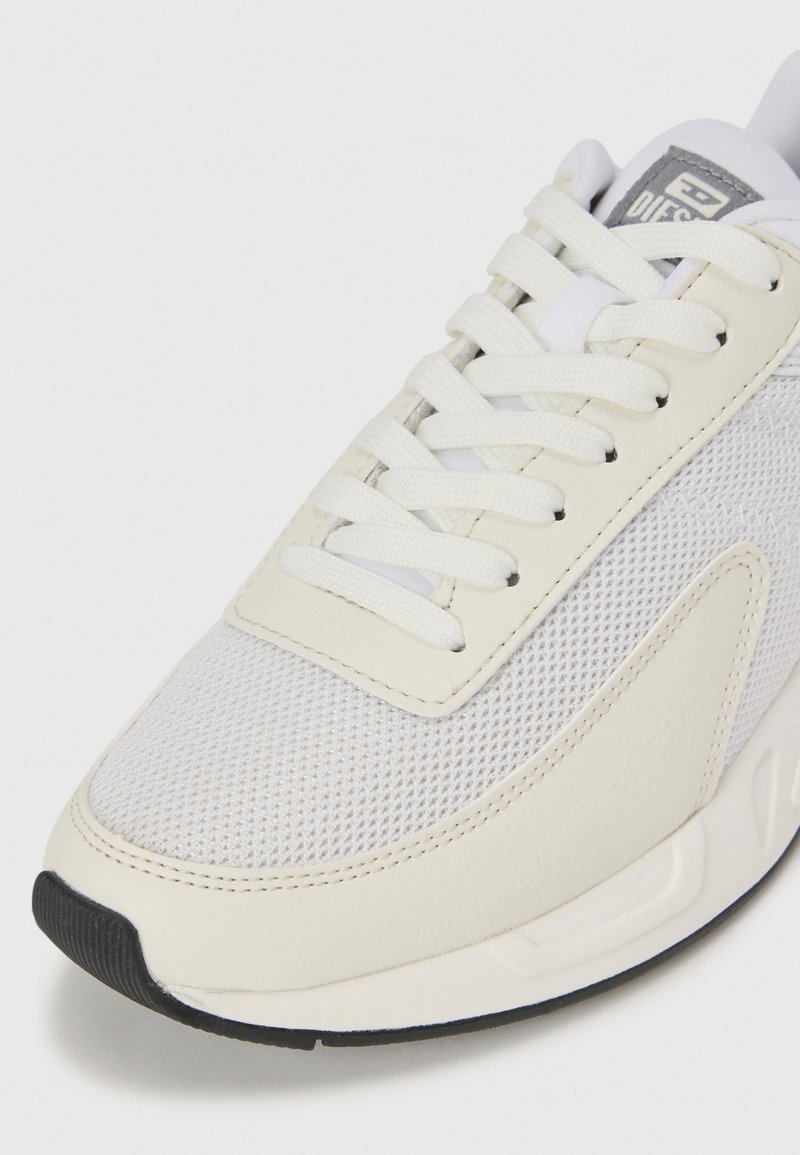 Sneaker blanche avec un dessus en maille texturée, un revêtement en cuir lisse, des lacets plats, et une semelle en caoutchouc noire contrastante. Accents gris clair.