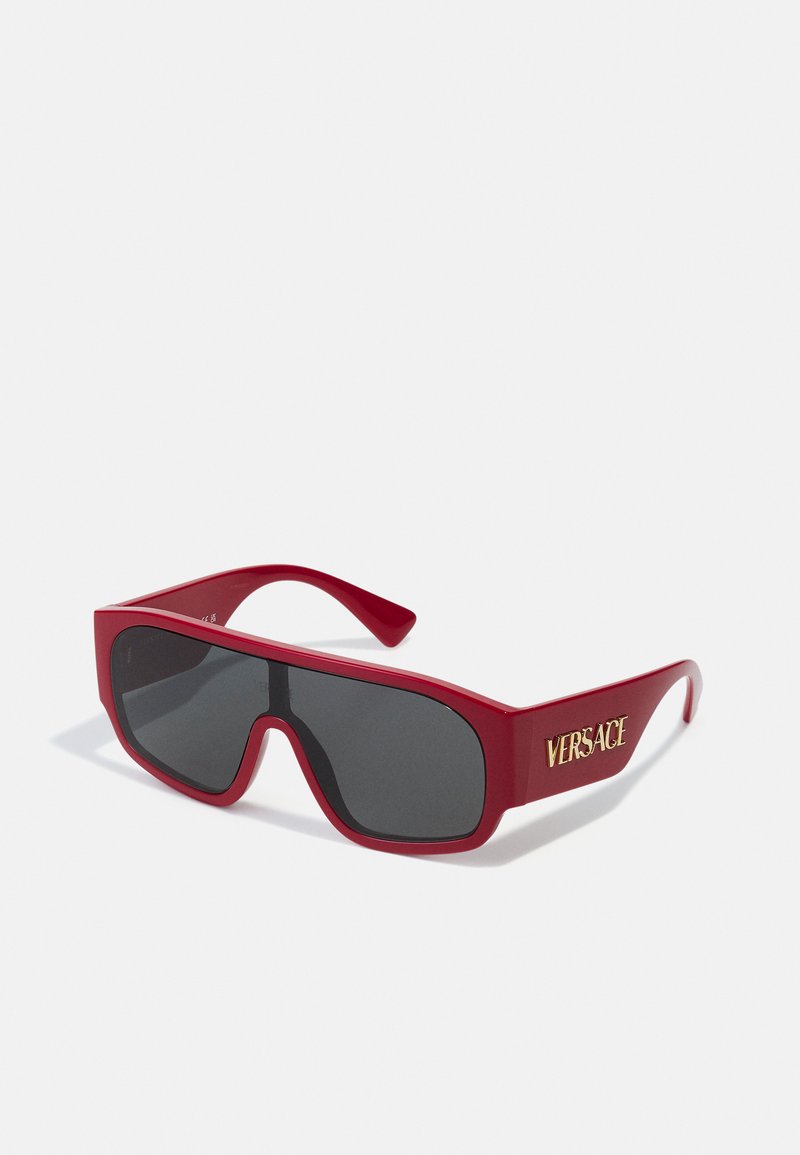 Ochelari de soare din plastic roșu, cu o formă largă, care se înfășoară în jurul feței și cu lentile negre. Prezintă un logo auriu "VERSACE" pe brat. Textură netedă.