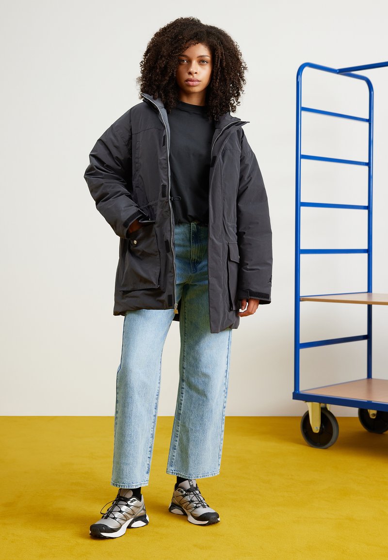 Jeune femme aux cheveux bouclés portant une veste noire oversize, une chemise noire, un jean bleu clair et des baskets noires et argentées, se tenant sur un sol jaune.