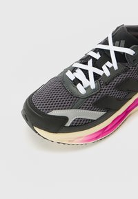 Black and gray athletic shoe with a mesh upper, white laces, silver accents, and a pink-to-beige gradient sole. Features textured materials.