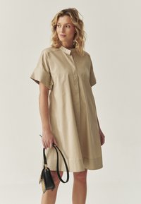 Beige, lyhythihaine mekko napilla varustetulla kauluksella ja rentoa istuvuutta, jossa on sileä pinta. Malli pitää pientä mustaa laukkua, jossa on hihna.