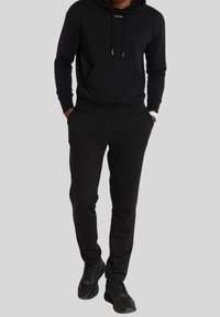 Uomo che indossa un hoodie nero di Calvin Klein, pantaloni jogger neri, sneakers nere e un orologio bianco, in piedi con le mani in tasca.