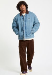 Lichtblauwe denimjas met een bruine corduroy kraag, drukknoopsluitingen, twee voorkruimelzakken, gecombineerd met bruine corduroy broeken en paarse sneakers.
