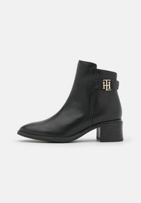 Bottine en cuir noir avec un bout pointu, un talon en bloc de hauteur moyenne et des accents en métal doré. Texture lisse avec un minimalisme dans les détails.