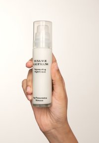 Bouteille cylindrique blanche avec un distributeur pompe, un capuchon transparent et une étiquette en texte noir indiquant "Crème de nuit régénérante" et "Soins préventifs contre le vieillissement."