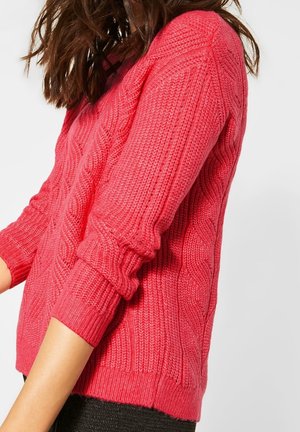 Kvinde iført en lys pink kabelstrikket sweater med opkrogede ærmer foran en ensartet lys baggrund.