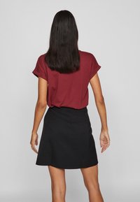 Μπορντό κοντομάνικο μπλουζάκι που συνδυάζεται με μαύρη φούστα που φουσκώνει, εικονιζόμενο από πίσω. Το ύφασμα φαίνεται λείο με κλασική σιλουέτα.