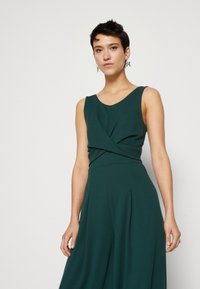 Bodysuit sans manches de couleur vert foncé avec un design de buste torsadé, jambes évasées et texture de tissu lisse. Il présente un décolleté en V et des plis.